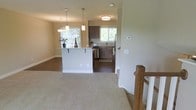 This two-story home offers a thoughtfully designed main level centered around an open-concept layout that connects the living area and kitchen. A carpeted living space transitions into the kitchen zone via hardwood-style dark flooring, creating a clear yet fluid distinction between areas. The kitchen features dark wood cabinetry paired with light stone countertops, a stainless steel appliance, and a peninsula with bar seating — ideal for casual dining or entertaining. Pendant lighting above the peninsula adds a stylish focal point, while a flush-mount ceiling fixture illuminates the kitchen proper. A window above the sink and an additional window near the dining area invite natural light and views of surrounding greenery, enhancing the airy feel of the space. Neutral-toned walls throughout contribute to a clean, cohesive aesthetic. A staircase with wooden balusters and white spindles connects the main level to the second floor, adding a classic architectural detail. The overall ambiance is warm, functional, and well-lit, making the main level both practical and inviting.