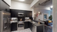 Located at 1601 Broadway in San Diego, unit #204 is a single-floor loft-style space defined by its industrial architectural character. Exposed concrete ceilings with visible structural beams and robust concrete columns run throughout, creating a raw yet refined urban aesthetic. Polished concrete floors extend across the living and entry areas, reinforcing the industrial design language.

The kitchen is a standout feature, outfitted with matte black cabinetry, dark subway tile backsplash, and a full suite of stainless steel appliances including a gas range, built-in microwave, side-by-side refrigerator, and dishwasher. Gray stone countertops and patterned tile flooring complete the space, while pendant lighting adds a warm, curated touch overhead.

The living area is generous and open, anchored by large windows that overlook a lush, tropical courtyard garden, flooding the space with natural light and greenery views. A glass door provides direct access to the outdoor area. Exposed structural columns divide the space organically, adding architectural interest.

The bedroom features a dramatic textured wood accent wall as a focal point, complemented by natural light from floor-to-ceiling glazing. The overall ambiance balances urban industrial elements with warmth, making this a distinctive and visually compelling urban residence.