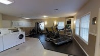 This single-floor fitness space offers a practical and well-organized environment for exercise and laundry needs. The room features warm beige walls accented by a white chair rail running the length of the space, complemented by white-trimmed windows fitted with slatted blinds that allow natural light to filter in, creating a bright and welcoming atmosphere.

The exercise area is anchored by rubberized flooring mats that delineate the workout zone and protect the light-toned hard floor beneath. Cardio equipment includes a treadmill and an elliptical machine, both positioned toward the windowed wall. A wall-mounted flat-screen television is conveniently placed nearby, allowing users to stay entertained during workouts. A multi-function cable strength training station is positioned toward the back of the room, accompanied by a workout bench, providing options for a range of resistance exercises.

Along the left wall, a set of overhead cabinets provides storage, and a washer is visible, indicating an in-unit laundry area integrated into the space. Ceiling-mounted light fixtures and wall sconces ensure adequate illumination throughout. Overall, the space is functional, cleanly designed, and efficiently organized.