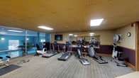 This fitness center offers a practical and well-organized space designed to support a variety of cardio-focused workouts. The room is furnished with multiple treadmills, elliptical trainers, and a recumbent bike, providing users with a solid range of exercise options. A large wall mirror runs along one side of the room, visually expanding the space and allowing users to monitor their form during workouts. A flat-screen television is mounted on the wall, adding an entertainment element to exercise sessions. The flooring features a striped, low-pile carpet that adds texture and warmth to the space. Overhead, a drop ceiling with recessed panel lighting provides even, consistent illumination throughout the room. A deep burgundy wainscoting accent runs along the lower portion of the walls, adding a touch of color contrast. Glass partitioning along one side of the room offers a view into what appears to be an adjacent indoor pool area, enhancing the sense of openness and connectivity to other amenities. The overall ambiance is functional and clean, making it a convenient option for those seeking an accessible, no-frills fitness facility.