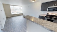 Located at 4351 15th Ave S in Seattle's South Seattle neighborhood, Unit 309 is a thoughtfully designed single-floor residence that combines modern aesthetics with functional living. The open-concept layout connects the kitchen and living area seamlessly, creating an inviting and spacious atmosphere.

The kitchen is outfitted with sleek dark cabinetry featuring brushed metal hardware and a generous stone countertop with warm, natural veining — a striking centerpiece that extends into a peninsula, offering additional prep space and the potential for casual dining or entertaining. The countertops continue onto the adjacent surfaces, providing ample workspace and visual continuity throughout.

The living area benefits from a large window that fills the space with natural light and frames views of the surrounding neighborhood and greenery. The light wood-toned laminate flooring runs throughout, contributing to a clean, contemporary feel while remaining durable and easy to maintain.

Crisp white walls and recessed flush lighting complete the interior, lending the unit a bright, neutral palette that adapts easily to a variety of furnishing styles. The overall design balances modern finishes with practical functionality, making this unit a comfortable and stylish residence in Seattle.
