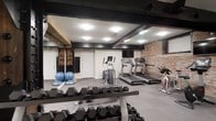 This fitness space combines industrial character with practical functionality, creating an inviting environment for a comprehensive workout. The room is defined by its exposed brick accent wall, which adds a raw, textural quality to the space, contrasting with the clean white walls on the opposite side. Dark-painted ceiling beams run overhead, contributing to the room's distinctive aesthetic while reinforcing its industrial character. The flooring consists of durable rubber gym matting, providing safety and comfort during exercise. A full-length mirror along one wall enhances the sense of space and allows users to monitor their form. The cardio section is well-appointed, featuring multiple treadmills, an elliptical machine, and a stationary spin bike, all positioned for easy access. A wall-mounted flat-screen television keeps exercisers entertained during cardio sessions. The free weights area includes a fully stocked dumbbell rack, offering a wide range of weight options. Stability and balance tools, including medicine balls, are also present. Overhead flush-mount lighting ensures the space is well-lit throughout, creating a bright and functional workout environment.