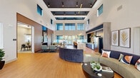 Located at 4525 Lacuna Ln in Kissimmee, Florida, this single-floor community clubhouse at The On Vine Apartment Homes offers a generously sized, thoughtfully designed gathering space. The centerpiece is a dramatic open living area with soaring double-height ceilings, clerestory windows, and an abundance of natural light that floods the warm hardwood-style flooring throughout. Large windows and glass doors frame views of the surrounding lush Florida landscape, including palm trees and a resort-style pool deck, which is accessible directly from the main living space. The main lounge is furnished with curved sectional seating, a bold round coffee table, and curated wall art, creating a welcoming and social atmosphere. A sleek kitchen bar with navy cabinetry, a stone backsplash, pendant lighting, and bar-height seating serves as both a functional and stylish focal point. A casual dining area with eclectic multi-colored chairs and bistro-style tables is positioned near the pool-facing glass doors. A separate, more intimate meeting or lounge room features a distinctive patterned accent wall, contemporary artwork, and flexible seating. The corridor areas are finished with warm wood-trimmed doorways, light neutral walls, and carpet-accented flooring, connecting the various zones of the space efficiently and elegantly.