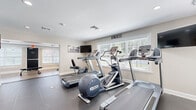 Located at 6139 Edward St in Norfolk, VA, this fitness center is a thoughtfully designed single-floor facility offering a comprehensive workout environment. The space is organized into distinct functional zones, providing ample room for a variety of exercise routines.

The main cardio area is outfitted with multiple Precor treadmills, an elliptical trainer, and a recumbent bike, all positioned near large windows fitted with white blinds that allow generous natural light to fill the room. Wall-mounted flat-screen televisions are strategically placed throughout to keep users entertained during workouts. Motivational wall art, including a "Feel the Burn" sign, adds an energetic atmosphere to the space.

A dedicated strength training area houses a multi-function cable machine and a flat weight bench, while a separate free weights section features a tiered dumbbell rack stocked with a full range of weights. This area is finished with light wood-look flooring and accented by a textured brick-style wall and a large floor-to-ceiling mirror that visually expands the space. Colorful exercise balls on a storage rack add a vibrant touch.

The flooring throughout transitions between durable rubber gym matting and smooth hard flooring, suited to each area's function. Recessed lighting and standard overhead fixtures ensure consistent illumination. The facility is accessed via a glass exterior door, with a clearly marked emergency exit for safety.