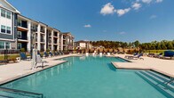 Located at 1130 Annondale Way in Fayetteville, North Carolina, this outdoor amenity space delivers a resort-style experience designed for relaxation and recreation. The centerpiece is a generously sized swimming pool with crystal-clear blue water, surrounded by a broad, well-maintained concrete deck lined with blue lounge chairs and chaise seating — ideal for sunbathing under the North Carolina sky. A second, smaller pool and a dedicated spa area provide additional aquatic options, catering to a range of preferences. Beyond the pools, a spacious lounge zone features comfortable outdoor sectional sofas and chairs, offering a comfortable setting for socializing or unwinding. An outdoor grilling station with multiple gas grills and adjacent dining tables provides a convenient space for outdoor cooking and dining. The perimeter is enclosed by a sleek black iron fence, with landscaped greenery and mature trees offering a natural backdrop. Lush open lawn areas extend beyond the pool deck, adding to the sense of openness. The multi-story residential buildings surrounding the space feature contemporary architecture with mixed siding, balconies, and large windows. The entire amenity area is well-appointed, open to abundant natural light, and designed to complement an active outdoor lifestyle.