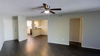 This single-story space in Irving, Texas, offers an open and functional layout designed for comfortable everyday living. The main living area features rich, dark hardwood-style flooring that contrasts cleanly with the neutral beige walls and bright white baseboards, creating a polished, cohesive aesthetic throughout. A ceiling fan with a built-in light fixture is centrally mounted on the textured ceiling, providing both airflow and ambient lighting to the space.

The living area flows seamlessly into the kitchen, which is visible through an open pass-through arrangement. The kitchen is equipped with white cabinetry, a stainless steel range, and a breakfast bar that offers additional seating and countertop space. HVAC vents are positioned throughout the ceiling, ensuring efficient climate control across the open floor plan.

A doorway off the main living area leads to additional rooms, suggesting a practical and well-connected layout. The overall ambiance is clean, bright, and move-in ready, with a neutral color palette that provides a versatile backdrop suitable for a variety of furnishing styles. The single-floor design ensures easy accessibility and a straightforward flow between living spaces.
