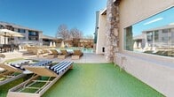 Located at 5700 Market St in Prescott Valley, Arizona, this outdoor amenity space is thoughtfully designed for communal gathering and relaxation. The centerpiece of the area is a sparkling swimming pool surrounded by a generous concrete deck lined with cushioned lounge chairs and striped sun loungers, ideal for soaking in the abundant Arizona sunshine. Artificial turf sections provide a clean, low-maintenance ground covering throughout the space, adding a touch of greenery year-round. A standout feature is the substantial stone-clad fire pit, constructed with stacked stone detailing and a decorative glass or rock fill, flanked by seating for intimate evening gatherings. An outdoor kitchen area includes multiple stainless steel built-in grills set into a stacked stone surround, making it well-suited for social events and barbecues. A covered patio section with tile flooring and wall-mounted lighting offers a shaded retreat, accessible through elegant white French doors with glass panels that connect to an interior space. Natural stone accent walls lend a rugged, Southwestern character to the architecture, complementing the neutral stucco exterior. The entire area enjoys open views of the surrounding neighborhood and clear blue skies, creating a bright and inviting outdoor environment.
