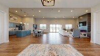 This single-story community facility is thoughtfully designed to serve as a versatile gathering and amenity hub. The welcoming entryway greets visitors with warm wood-look flooring, a patterned area rug, and a pair of accent chairs positioned beside a modern glass-panel entry door, with natural light filtering through blinds.

The heart of the space is a generously proportioned open-concept great room that seamlessly connects the kitchen, dining, and lounge areas. Vaulted ceilings rise over the lounge, which centers on a sleek electric fireplace set into a textured tile surround beneath a wall-mounted television. Sliding glass doors lead to an outdoor patio, filling the area with abundant natural light. A statement chandelier anchors the central gathering zone, while a large dining table accommodates group seating.

The kitchen features bold blue-gray cabinetry, marble-look countertops, a substantial center island with an undermount sink, stainless steel appliances, and dramatic pendant lighting above soaring ceilings.

A dedicated fitness room houses cardio and strength equipment alongside a wall-mounted television and wood-look flooring. The office area provides conference-style seating, individual workstations, and natural light through large windows, offering a functional workspace within the facility.
