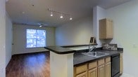 This single-floor residence offers an open-concept layout that seamlessly connects the kitchen and living area, creating a spacious and functional flow throughout the space. The kitchen is outfitted with light wood cabinetry, dark speckled countertops, and a stainless steel sink, complemented by a peninsula that extends the counter space and serves as a natural dividing element between the kitchen and living area. Track lighting mounted on the ceiling provides focused illumination throughout the kitchen, while a ceiling fan with a light fixture keeps the living area comfortable and well-lit. Rich hardwood flooring runs throughout the entire space, adding warmth and a cohesive aesthetic. A large window in the living area frames views of lush greenery outside, allowing abundant natural light to fill the room and creating a bright, airy ambiance. The clean, neutral wall tones throughout the space enhance the sense of openness, making it feel welcoming and versatile. The overall design balances practicality with comfort, offering an inviting environment ideal for everyday living and entertaining.