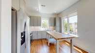 This modern single-floor residence showcases a clean, contemporary design with warm wood-tone laminate flooring that flows consistently throughout the entire space, tying each room together cohesively. The open-concept kitchen is a standout feature, outfitted with sage-gray shaker-style cabinetry, white quartz countertops, a classic subway tile backsplash, and stainless steel Whirlpool appliances including a range, over-the-range microwave, and refrigerator. A freestanding butcher block island provides additional workspace and adds a natural wood accent to the kitchen. Multiple windows flood the kitchen with natural light and offer views of surrounding greenery. The adjacent living area is bright and airy, benefiting from large windows and a sliding glass door that opens to a private balcony with white railings, extending the living space outdoors. The bedroom is a comfortable, light-filled room with a window overlooking trees, recessed lighting, and ample space. The bathroom is thoughtfully designed with gray cabinetry, white quartz countertops, a wide vanity mirror with overhead lighting, and a frameless sliding glass walk-in shower with white tile surround. Recessed lighting and roller shades are found throughout, contributing to the space's clean, modern aesthetic.