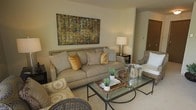 This single-floor residence showcases a carefully curated living space that balances comfort and style. The living area serves as the focal point of the home, featuring a large, plush neutral-toned sofa accented with golden and cream decorative pillows that introduce warmth to the overall palette. A bold, abstract canvas artwork anchors the main wall, adding a rich mix of earthy tones—greens, golds, and browns—that tie the room's color scheme together cohesively.

A glass-topped coffee table with a metal frame occupies the center of the seating area, styled with decorative trays, woven baskets, and small potted greenery, adding texture and a natural element to the space. Wicker accent chairs complement the neutral furnishings, reinforcing a relaxed yet refined aesthetic. A classic table lamp on a dark side table provides warm ambient lighting.

The space flows with a consistent design language—light walls, neutral carpeting, and warm wood tones visible in doorways contribute to a calm, welcoming atmosphere. Overall, the residence presents a harmonious and functional layout with tasteful décor that creates an immediately comfortable and livable environment.