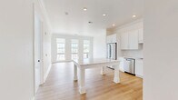 Located at 1 First St in Oakmont, PA, this well-appointed one-bedroom, one-bathroom residence offers a thoughtfully designed single-floor layout. The open-concept kitchen and living area serves as the heart of the home, featuring light hardwood flooring, crown molding, and recessed lighting throughout. The kitchen is outfitted with crisp white shaker-style cabinetry, a white subway tile backsplash, stainless steel appliances including a French door refrigerator, range, microwave, and dishwasher, and a standout white island with elegant column legs and a light stone countertop. A wall of tall windows in the living area floods the space with natural light and frames views of the surrounding Oakmont neighborhood. The bedroom offers plush carpeting, a sliding glass door opening to a private balcony with outdoor views, and a closet with clean white panel doors. A hallway connects the main living spaces to the full bathroom, which features earthy tile flooring, a white vanity with a large mirror, and a glass-enclosed walk-in shower. The overall aesthetic is clean, modern, and move-in ready, with a bright, airy ambiance maintained consistently throughout the home.