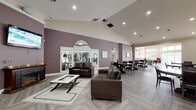 This single-story community clubhouse is thoughtfully designed to offer a variety of social, recreational, and functional spaces. The entry foyer sets an inviting tone with its herringbone-patterned flooring, vaulted ceiling, arched transom entry doors, and decorative wall accents. The main lounge area features soaring vaulted ceilings, warm mauve accent walls, a fireplace with a mounted flat-screen TV, and rich leather seating arranged over a herringbone-tiled floor. Large arched windows provide views directly into the adjacent fitness center, which is equipped with a range of strength and cardio machines. The dining area offers multiple seating arrangements, including a round glass-top table and counter-height seating, all bathed in natural light from expansive windows and glass doors leading to an outdoor area. A dedicated billiards room features rich wood wainscoting, a full-size pool table, a wall-mounted TV, and a window overlooking the outdoors. The conference room impresses with a tray ceiling in deep mauve, recessed lighting, a tile-surround fireplace with a mounted TV, built-in white bookshelves, French doors to the exterior, and a large oval conference table surrounded by tufted leather chairs—ideal for formal meetings or private gatherings.