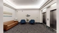 Located at 201 Broad Street in Stamford, Connecticut, this building lobby offers a refined and welcoming first impression. The entryway features striking dark green marble tile flooring with white veining, which transitions into patterned carpeting throughout the main seating area. Glass-paneled entry doors allow natural light to filter into the vestibule, creating a bright and accessible entry point.

The lobby's seating area is thoughtfully furnished with a tan leather sofa flanked by mirrored side tables and silver glass-base lamps, complemented by a pair of tufted navy blue accent chairs arranged around a gold geometric side table adorned with a fresh floral arrangement. Above the chairs, a decorative cluster of circular gold-framed mirrors serves as a focal point on the neutral gray wall. An abstract canvas painting hangs above the sofa, adding a touch of contemporary art to the space.

Recessed tray ceiling detailing with integrated ambient lighting lends the lobby a polished, modern aesthetic. A bank of two stainless steel elevator doors provides vertical access throughout the building, finished with sleek brushed metal panels and green indicator displays. The overall ambiance is professional yet comfortable, reflecting a well-maintained and thoughtfully designed commercial building lobby.