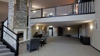 This expansive common area showcases a striking double-height ceiling that creates an open, airy atmosphere. A prominent stone-clad fireplace anchors one side of the space, adding warmth and a classic architectural detail. Comfortable seating, including upholstered armchairs arranged around a small table, provides a welcoming lounge area for residents or guests.

An upper mezzanine level overlooks the main floor, enclosed by a dark-railed balustrade with contrasting white spindles, adding visual depth to the vertical space. Wall-mounted sconces provide warm, ambient lighting throughout. A reception or service counter is positioned nearby, alongside elevator access that connects the floors conveniently.

The flooring features neutral-toned carpet that extends across the main level, complementing the taupe and grey wall tones. The overall design balances functional layout with inviting aesthetics, reflecting a well-appointed communal environment suited to a residential building or boutique hospitality setting.