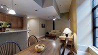 This single-floor residence features an open-concept layout that seamlessly connects the dining and living areas, creating a cohesive and functional living environment. The rich hardwood flooring extends throughout the main space, adding warmth and visual continuity. The dining area is anchored by a classic wooden table with Windsor-style chairs, complemented by a decorative bowl centerpiece. A peninsula counter separates the kitchen from the dining space, offering additional functionality.

The living area is comfortably appointed with a neutral-toned sofa, accent armchair, and a wooden coffee table set atop a area rug. A ceiling fan with integrated lighting serves as a practical overhead fixture. The walls are finished in warm taupe tones, contributing to the overall inviting ambiance. Recessed lighting and a framed wall piece add subtle decorative touches.

A dedicated laundry area, visible through a doorway off the kitchen, houses stacked washer and dryer units, providing convenient in-unit laundry access. The space benefits from varied ceiling heights, with architectural transitions adding visual interest overhead.