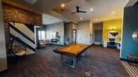 Located in Fort Worth, TX, this single-floor amenity space blends industrial character with contemporary design across several distinct activity zones. Exposed brick walls, patterned carpeting, and bold accent colors create a dynamic, urban atmosphere throughout.

The game room anchors the space with a full-sized pool table beneath a ceiling fan and recessed lighting, flanked by a lounge area with a sofa, fireplace, and sputnik-style chandelier. Nearby, stylish egg-shaped hanging chairs add a playful touch alongside vibrant wall art and a bold diagonal-striped accent wall.

The kitchen features deep navy cabinetry, stainless steel appliances, a striking geometric-patterned island with bar seating, and exposed brick as a dramatic backdrop, all illuminated by Edison-style pendant lighting. Adjacent dining tables with industrial metal stools and a wall-mounted TV offer a collaborative gathering spot, complemented by a large cactus mural.

The lounge area is lined with panoramic windows and a built-in window bench, offering sweeping views of the Fort Worth skyline and surrounding cityscape. Plush swivel chairs, a ceiling fan, and access to an outdoor balcony make this a bright, inviting retreat. A dedicated workspace with a desk and office chair rounds out this well-appointed community space.
