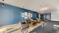 The lobby at 4875 Dufferin Rd in Montréal, QC serves as an elegant and well-designed entrance that sets a refined tone for the building. The most striking architectural feature is the boldly panelled accent wall finished in a deep steel blue, which spans the length of the seating area and immediately draws the eye upon entry. Decorative moulding frames each panel, lending a classic yet contemporary character to the space.

The flooring transitions between polished terrazzo tiles — adorned with an intricate inlaid mosaic pattern in warm terracotta, white, and dark green tones — and grey carpeted areas that define the circulation paths leading to the elevators and staircase. This contrast adds visual depth and clearly delineates zones within the lobby.

The seating area is furnished with a taupe leather sofa and matching armchairs arranged around a gold-accented wood coffee table, creating an inviting lounge setting. Wall-mounted sconces and pendant lighting throughout provide warm, layered illumination, complemented by a chandelier near the elevator bank. Curated abstract artwork and a potted tropical plant add personality to the space.

A glazed leasing office with frosted signage is accessible directly from the lobby, and two elevators framed by grey stone-tiled surrounds provide vertical access to upper floors. The overall ambiance is polished, welcoming, and thoughtfully appointed.
