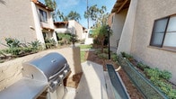 This single-story outdoor common area showcases classic Spanish Colonial-inspired architecture, characterized by warm stucco exteriors, red clay tile rooflines, and exposed wooden eave detailing. The buildings surrounding the courtyard feature neutral tan stucco walls with brown-trimmed windows, creating a cohesive and inviting aesthetic throughout the complex.

The communal outdoor space is well-appointed with practical amenities, including a full-size stainless steel gas grill station ideal for outdoor entertaining and social gatherings. Adjacent to the grill area, metal bench seating provides comfortable spots for residents to relax and enjoy the surroundings. Classic black lamp posts line the concrete pathways, adding a decorative touch while ensuring the space remains well-lit.

The landscaping throughout the courtyard is lush and thoughtfully designed, featuring tall swaying palm trees, eucalyptus trees, bird-of-paradise plants, and a variety of tropical and drought-tolerant shrubs and ground cover. Neatly maintained green lawns and mulched garden beds wind between the buildings, contributing to an overall resort-like atmosphere. The open layout and sunny California sky visible above create a bright, welcoming outdoor environment suited for year-round enjoyment.