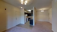 This single-floor unit presents an open-concept design that seamlessly connects the kitchen and living areas, creating a cohesive and functional layout. The kitchen is equipped with white appliances, including a refrigerator and range, and is complemented by dark cabinetry that provides a strong visual contrast against the light-colored walls. A tile floor grounds the kitchen space, while a peninsula with dark cabinetry serves as a natural divider between the kitchen and the adjacent dining or living area. The living area features soft carpeting and is illuminated by a ceiling fan with an integrated light fixture, adding both practicality and a touch of style. A secondary chandelier-style light fixture is visible in the adjoining space, enhancing the overall brightness of the unit. The front entry door is positioned conveniently near the kitchen, offering direct access to the main living space. Neutral wall tones throughout create a clean, understated backdrop that allows for flexible interior styling. The layout is efficient and well-organized, making the most of the available space.