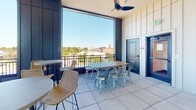 Located at 143 E White St in Rock Hill, South Carolina, this single-level rooftop terrace offers a well-appointed open-air gathering space with a refined, contemporary design. The space is defined by bold navy-blue paneled walls and structural columns that frame expansive floor-to-ceiling windows and open railings, flooding the area with natural light and providing panoramic views of the surrounding downtown Rock Hill cityscape.

The terrace is furnished for versatile use, featuring a mix of high-top bistro tables with woven rattan bar chairs and a longer communal dining table paired with industrial-style metal seating—ideal for both casual mingling and group gatherings. Ceiling fans are mounted overhead to ensure comfort during warmer months.

A striking focal point of the space is a floor-to-ceiling fireplace clad in layered brick and glossy dark tile, accented by a warm wood mantel and flanked by built-in bench seating with decorative cushions, creating an inviting lounge area.

A built-in wet bar with a stainless steel countertop, undermount sink, under-counter refrigeration, and a white tile backsplash illuminated by vintage-style sconce lighting adds a functional and stylish amenity to the space.