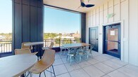 Located at 143 E White St in Rock Hill, South Carolina, this single-level rooftop terrace offers a well-appointed open-air gathering space with a refined, contemporary design. The space is defined by bold navy-blue paneled walls and structural columns that frame expansive floor-to-ceiling windows and open railings, flooding the area with natural light and providing panoramic views of the surrounding downtown Rock Hill cityscape.

The terrace is furnished for versatile use, featuring a mix of high-top bistro tables with woven rattan bar chairs and a longer communal dining table paired with industrial-style metal seating—ideal for both casual mingling and group gatherings. Ceiling fans are mounted overhead to ensure comfort during warmer months.

A striking focal point of the space is a floor-to-ceiling fireplace clad in layered brick and glossy dark tile, accented by a warm wood mantel and flanked by built-in bench seating with decorative cushions, creating an inviting lounge area.

A built-in wet bar with a stainless steel countertop, undermount sink, under-counter refrigeration, and a white tile backsplash illuminated by vintage-style sconce lighting adds a functional and stylish amenity to the space.