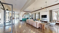 This expansive single-floor clubhouse is defined by its refined architectural design, blending classical and contemporary elements throughout. The grand entrance lobby sets the tone immediately, featuring soaring ceilings, bold white structural columns, rich wood-stained exposed beams, and large-format tile flooring with striking dark inlay accents. Walls of black-framed glass allow abundant natural light to pour in while framing views of the outdoor pool and surrounding grounds.

The open lounge areas are anchored by warm hardwood-style flooring, elegant crown molding with dark wood trim, and statement chandelier lighting. Arched doorways and arched glass windows create graceful transitions between zones, contributing to a cohesive and inviting atmosphere. Seating areas are generously appointed, with ample space for both relaxed gathering and quiet retreat.

A dedicated games area features a full-size pool table beneath exposed wood beams, with floor-to-ceiling glass walls opening toward the pool terrace. A private workstation area showcases a bold geometric paneled accent wall in deep gray, housing dual desks for focused productivity. A bright conference or dining room offers direct pool views through a dramatic arched floor-to-ceiling glass facade, completing this well-appointed communal facility.