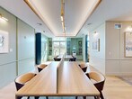 This library and co-working space in Peabody, Massachusetts offers a refined, multi-functional environment designed to support both focused individual work and collaborative study. The central library area is anchored by long, light wood communal tables paired with stylish mid-century-inspired chairs, illuminated by sleek gold pendant lights suspended from a coffered ceiling with warm wood trim detailing. The walls are dressed in soft sage green tones, accented by deeper teal cabinetry and built-in bookshelves that line the far wall, adding both functionality and visual depth. Natural light pours in through large, grid-paned windows, offering views of the surrounding greenery and creating a bright, welcoming atmosphere. A dedicated work pod area provides semi-private workstations equipped with height-adjustable desks, mobile filing cabinets, and comfortable seating — ideal for focused, heads-down tasks. Tucked away from the main areas, intimate reading nooks feature plush leather armchairs, ottomans, accent tables, and elegant wall sconces, offering a quiet retreat for leisure reading or private reflection. Light hardwood flooring flows throughout, complemented by textured area rugs that define each zone. The overall ambiance is calm, sophisticated, and highly functional.