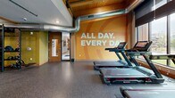 Located at 4043 Chatsworth St N in Shoreview, Minnesota, this fitness center offers a generously sized, single-floor exercise facility designed to support a full range of workout activities. The space is divided into distinct functional zones, each thoughtfully appointed to serve different training needs.

The cardio area features multiple treadmills, elliptical machines, and a StairMaster, positioned in front of expansive floor-to-ceiling windows with roller shades. These large windows invite abundant natural light and provide open views of the surrounding greenery, creating an energizing environment. Exposed wood beams and visible HVAC ductwork lend an industrial-contemporary aesthetic to the ceiling.

A dedicated studio zone includes hardwood flooring, floor-to-ceiling mirrors, and ample open space suitable for cycling classes, yoga, or group fitness. Stationary bikes are neatly arranged near large windows, maximizing both light and ventilation.

The strength and functional training area is outfitted with multi-tier equipment racks stocked with medicine balls, kettlebells, and resistance tools, as well as cable machines and weight benches on durable rubberized flooring.

The entrance area makes a bold impression with motivational wall graphics and warm wood accents. Decorative plants add a welcoming touch. Recessed lighting throughout ensures consistent illumination across all zones, rounding out a professional, well-maintained fitness environment.