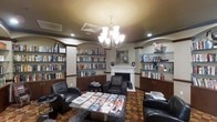 This single-floor library lounge is designed as a comfortable and sophisticated reading space. Built-in bookshelves line the walls on both sides of the room, filled with an extensive collection of books and media, framed by arched alcoves that add architectural character. The shelves are complemented by dark wood cabinetry below, providing additional storage while reinforcing the room's refined aesthetic.

A white-mantled fireplace serves as the room's focal point, anchored by a framed portrait hung above it, adding a classic and elegant touch. The space is illuminated by a multi-arm chandelier suspended from the ceiling, supplemented by recessed lighting that ensures a warm, well-lit ambiance throughout.

Seating is arranged thoughtfully around a central coffee table, with dark leather armchairs offering comfortable spots for reading or relaxing. The table displays an assortment of magazines, making the space feel both functional and welcoming. Underfoot, a bold patterned carpet adds visual interest and warmth to the room. Overall, the space balances a classic library atmosphere with comfortable, lounge-style furnishings.