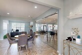 Located at 5080 Samet Dr in High Point, NC, this well-appointed community clubhouse offers a thoughtfully designed single-floor layout encompassing a living room, dining room, and kitchen. The interior is finished with soft blue-gray walls and warm wood-look herringbone tile flooring throughout the main areas, creating a cohesive and welcoming atmosphere. A striking full-height glass sliding wall separates the dining area from the adjoining lounge space, maintaining an open feel while offering flexible room configurations. The kitchen features a curved white island with bar seating, geometric brass pendant lighting, white cabinetry, and a decorative mosaic tile backsplash. The living room benefits from vaulted ceilings, abundant natural light from large multi-pane windows, and a comfortable seating arrangement centered around a media console. A private office with carpeted flooring, recessed lighting, and a large window provides a functional workspace. The dollhouse view reveals a generous outdoor area complete with a resort-style pool, lounge seating, and a pergola structure, extending the usable space well beyond the interior footprint.