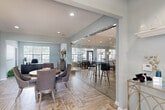 Located at 5080 Samet Dr in High Point, NC, this well-appointed community clubhouse offers a thoughtfully designed single-floor layout encompassing a living room, dining room, and kitchen. The interior is finished with soft blue-gray walls and warm wood-look herringbone tile flooring throughout the main areas, creating a cohesive and welcoming atmosphere. A striking full-height glass sliding wall separates the dining area from the adjoining lounge space, maintaining an open feel while offering flexible room configurations. The kitchen features a curved white island with bar seating, geometric brass pendant lighting, white cabinetry, and a decorative mosaic tile backsplash. The living room benefits from vaulted ceilings, abundant natural light from large multi-pane windows, and a comfortable seating arrangement centered around a media console. A private office with carpeted flooring, recessed lighting, and a large window provides a functional workspace. The dollhouse view reveals a generous outdoor area complete with a resort-style pool, lounge seating, and a pergola structure, extending the usable space well beyond the interior footprint.