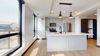 This contemporary single-floor residence showcases a thoughtfully designed open-concept layout with a clean, modern aesthetic throughout. The kitchen serves as a central highlight, featuring a large white quartz island with an undermount sink and matte black faucet, complemented by two striking conical pendant lights suspended from a recessed ceiling tray. Gray upper and lower cabinetry lines the walls, paired with stainless steel appliances including a gas range, built-in microwave, and French door refrigerator. Light hardwood flooring flows throughout the main living areas, enhancing the bright, airy atmosphere created by an abundance of large, black-framed windows.

A secondary galley-style kitchen configuration is also present, offering white cabinetry, matching quartz countertops, and city views through tall windows. The bedrooms feature plush carpeting and generous natural light, with one room showcasing an exposed concrete structural column — an industrial design detail that adds architectural character. A stylish barn-style sliding door adds functional flair to one of the rooms. The bathrooms are finished with white quartz double-vanity countertops, matte black hardware, and framed mirrors, while the half bath features a gray vanity and wood-look flooring.