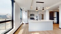 This contemporary single-floor residence showcases a thoughtfully designed open-concept layout with a clean, modern aesthetic throughout. The kitchen serves as a central highlight, featuring a large white quartz island with an undermount sink and matte black faucet, complemented by two striking conical pendant lights suspended from a recessed ceiling tray. Gray upper and lower cabinetry lines the walls, paired with stainless steel appliances including a gas range, built-in microwave, and French door refrigerator. Light hardwood flooring flows throughout the main living areas, enhancing the bright, airy atmosphere created by an abundance of large, black-framed windows.

A secondary galley-style kitchen configuration is also present, offering white cabinetry, matching quartz countertops, and city views through tall windows. The bedrooms feature plush carpeting and generous natural light, with one room showcasing an exposed concrete structural column — an industrial design detail that adds architectural character. A stylish barn-style sliding door adds functional flair to one of the rooms. The bathrooms are finished with white quartz double-vanity countertops, matte black hardware, and framed mirrors, while the half bath features a gray vanity and wood-look flooring.