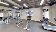 Park Hoover FC is a well-appointed fitness center designed to support a comprehensive workout experience. The facility features an exposed ceiling with industrial-style steel trusses and ductwork, lending the space a contemporary, open feel with generous overhead clearance throughout. Durable rubber flooring covers the workout areas, complemented by light wood-look flooring in the common and transition zones.

The cardio area is outfitted with treadmills, elliptical trainers, and stationary and spin bikes, positioned near large windows and glass doors that overlook an outdoor area, filling the space with natural light. A wall-mounted television provides entertainment during workouts.

The strength training section houses a collection of cable machines, weight benches, dumbbell racks, and a rowing machine. Floor-to-ceiling mirrors line the walls, enhancing the sense of space and allowing users to monitor their form. Decorative artwork adds a polished touch to the area.

A common area features bold blue accent walls, light wood flooring, a water fountain, and a vending machine, providing a functional transitional hub connecting the workout zones. Additional rooms off this area likely serve as restrooms or support spaces. The overall ambiance is clean, modern, and purpose-built for fitness.