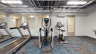 Located at 2525 E Main St in Richmond, Virginia, this fitness center offers a well-equipped, single-floor facility designed to support a comprehensive workout experience. The space features an open industrial aesthetic, highlighted by an exposed ceiling grid with integrated lighting that illuminates the entire floor plan clearly and efficiently.

The cardio zone is stocked with multiple treadmills, elliptical trainers, and a stationary bike, all arranged to allow for comfortable spacing between users. A dedicated strength training area includes a multi-function cable machine and a leg press/extension station, providing options for a full range of resistance exercises. Free weights are neatly organized on a tiered dumbbell rack along a mirrored wall, which also helps to visually expand the space.

Large windows along one wall allow natural light to filter into the facility, contributing to an energizing atmosphere. A water fountain station is conveniently mounted on the wall, and a sanitation dispenser ensures cleanliness throughout. A glass-paneled door leads to an adjacent room, offering additional functional space.

The flooring throughout consists of patterned modular carpet tiles in a blue and grey tone, adding a modern, polished feel to the environment. Overall, this fitness center is thoughtfully arranged to maximize usability while maintaining a clean, professional atmosphere.
