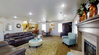 This single-floor residence offers a thoughtfully designed open-concept layout that seamlessly connects the living, dining, and kitchen areas into one cohesive and functional living space. The living room is generously proportioned, anchored by a large dark-wood entertainment center and complemented by a mix of comfortable seating, including a rich leather sofa and a powder-blue accent chair that adds a pop of color. A plush area rug and a round tufted ottoman sit at the center, enhancing the room's inviting atmosphere. A ceiling fan with integrated lighting keeps the space comfortable year-round. The dining area transitions naturally from the living room, offering space for a full dining table and chairs, ideal for everyday meals or entertaining. Adjacent to the dining space, the kitchen features light wood cabinetry, white appliances, and a breakfast bar with bar-height seating, providing additional casual dining options. Recessed lighting is installed throughout, ensuring consistent illumination across the open floor plan. Multiple windows flood the space with natural light, creating a bright and airy ambiance. The textured ceilings and neutral tones of the walls and carpeting provide a clean, classic backdrop throughout this well-appointed home.