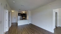 This single-floor space features an open-concept design that seamlessly connects the living area to the kitchen, creating a bright and functional layout. The flooring throughout is rich, dark hardwood-style plank, lending a contemporary and cohesive aesthetic to the space.

The kitchen is a standout feature, equipped with dark cabinetry that contrasts sharply against the white walls and light countertops. Stainless steel appliances, including a refrigerator, microwave, and range, are neatly integrated into the cabinetry. A peninsula with a granite-style countertop provides additional workspace and serves as a natural divider between the kitchen and the open living area. Pendant lighting with brass-toned fixtures hangs above the peninsula, adding a modern touch.

Adjacent to the kitchen, a built-in shelving unit is recessed into the wall, offering practical display or storage options. The living area is generously sized, with clean white walls and ample room for furnishings. A flush-mount ceiling fixture provides overhead lighting, while a visible HVAC vent ensures climate control. The entry door is positioned near the kitchen, featuring a modern smart lock for added security.