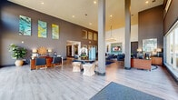 This expansive single-floor clubhouse offers a thoughtfully designed layout that blends functionality with contemporary style. The grand entrance lobby immediately impresses with soaring double-height ceilings, structural columns, and light wood-look flooring that flows throughout the space. Vibrant abstract artwork adorns the walls, while a central accent table with sculptural seating creates an inviting focal point. Adjacent to the entry, a stylish lounge area features comfortable navy seating and a wall-mounted display screen, ideal for relaxed social gatherings.

A private office off the lobby is bathed in natural light through floor-to-ceiling windows overlooking outdoor greenery, complemented by dramatic high ceilings and bold artwork above the executive desk. The open-plan kitchen and dining area showcases a large central island with a quartz countertop, oversized pendant lighting, and high bar seating, while interior glazed windows create visual connectivity to neighboring spaces.

The well-appointed fitness center spans two levels, with cardio equipment, free weights, and strength machines on the main floor, and additional equipment on an upper mezzanine level accessed by a staircase. Large exterior-facing windows flood the gym with natural light. Connecting hallways feature bold framed windows offering interior sightlines, reinforcing the open and interconnected character of the entire facility.