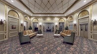Located at 5240 N Sheridan Road in Chicago's Edgewater neighborhood, this distinguished lobby exudes classical elegance through its richly detailed architectural design. The entrance corridor features a barrel-vaulted ceiling adorned with decorative gilded moldings and a chandelier, leading visitors across a polished marble floor laid in a geometric pattern to a staffed reception desk. Ornate wall sconces, framed artwork, and lush potted plants accent the passageway throughout.

The main lobby opens into a generously proportioned lounge area with a coffered ceiling trimmed in gold, walls adorned with intricate plasterwork, and grand arched transom panels featuring sunburst fan detailing. Warm, floral-patterned carpeting grounds the space, where seating arrangements of camel-toned sofas and wingback armchairs create comfortable, inviting conversation areas. Brass elevator doors with decorative inlays are framed by pilasters and ornamental moldings, accompanied by a coordinating marble-tiled elevator vestibule and a simple upholstered bench.

Throughout the lobby, the interplay of gilded accents, sculpted wall panels, classical column detailing, and carefully curated furnishings creates an atmosphere of timeless sophistication, evoking the grandeur of early 20th-century Chicago architecture.