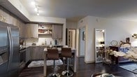 This single-floor residence showcases a thoughtfully designed open-concept layout that seamlessly connects the kitchen and living areas. The kitchen is a standout feature, appointed with contemporary taupe cabinetry, open display shelving, and a white subway tile backsplash that adds a clean, classic contrast. A granite-topped breakfast bar with room for two bar stools serves as both a functional dining space and a natural divider between the kitchen and the living area, making it ideal for casual dining or entertaining. Rich, dark hardwood flooring flows throughout, anchoring the space with warmth and sophistication. Track lighting illuminates the kitchen area, while the neutral wall tones create a bright and welcoming ambiance. A decorative full-length mirror with an ornate frame leans against the wall, adding depth and an elegant touch to the living space. A hallway visible from the main area leads to additional rooms, suggesting a well-organized and functional floor plan. Overall, the space balances modern design elements with comfortable, livable touches, resulting in a cohesive and stylish interior.
