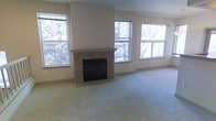 This single-floor living space offers a clean, functional layout centered around a standout tiled fireplace with a classic mantel surround. The fireplace serves as the focal point of the main living area, framed by neutral beige walls that create a warm, understated backdrop. The room is generously lit by multiple windows positioned on two walls, each offering views of mature trees outside, allowing abundant natural light to fill the space. The plush wall-to-wall carpeting in a neutral gray-green tone adds comfort underfoot and complements the overall soft color palette. The high placement of the windows, combined with the vaulted or elevated wall above the fireplace, contributes to an open, airy feel. The space is move-in ready with a clean, minimalist aesthetic, making it easy to personalize with furnishings of various styles. The combination of natural light, the warmth of the fireplace, and the soft carpeting gives the living area a cozy yet spacious ambiance well-suited for everyday living and relaxing.