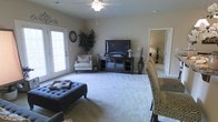 This single-story space centers around a well-appointed living room that exudes a comfortable, casual ambiance. The room is finished with plush, light-colored wall-to-wall carpeting that adds warmth underfoot, while neutral-toned walls create a versatile backdrop for a variety of décor styles. A ceiling fan anchors the ceiling, providing both function and a subtle design element.

Natural light is a notable feature, courtesy of French doors along one wall that offer a bright, airy feel to the interior. The living area is generously sized, accommodating a tufted ottoman serving as a central coffee table, a patterned accent chair, and a classic armchair tucked neatly in the corner beside a tall indoor plant. A dark wood media console holds a flat-screen television, completing the entertainment area.

Decorative wall accents and tall floor vases add character throughout the space. A doorway visible from the living room leads to an adjacent room, suggesting a connected and functional layout. Overall, the space presents a well-balanced blend of comfort and practicality, well-suited for everyday living.