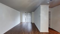 This single-floor space presents a clean, minimalist aesthetic defined by a few key architectural and design elements. The main living area is characterized by rich, dark hardwood flooring that contrasts sharply with the bright white walls, creating a visually striking and modern atmosphere. Crown molding runs along the upper edges of the walls, adding a subtle touch of classic detail to the otherwise contemporary space.

A full-width sliding glass door at the far end of the room serves as the focal point, fitted with vertical blinds that allow for adjustable natural light. This feature not only brightens the interior but also provides a visual connection to the outdoors. The walls are smooth and freshly painted in white, contributing to a bright and open feel throughout the space.

The room offers a generous, open layout with ample floor space, making it highly versatile for various furniture arrangements. Baseboards in white complement the overall clean design. The combination of the dark flooring, white surfaces, and natural light source gives the space a balanced, welcoming ambiance suitable for a range of uses.
