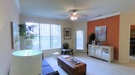 This single-floor residence features a welcoming living area designed with both comfort and style in mind. The room is anchored by a warm terracotta accent wall that adds a bold pop of color against the neutral, light-toned walls throughout. Crown molding adds a refined architectural detail along the ceiling line, while a ceiling fan with integrated lighting provides both function and a subtle decorative touch.

Natural light is a defining characteristic of the space, courtesy of large windows dressed with vertical blinds that allow for adjustable light control. A private exterior door with a windowed panel further brightens the room and provides convenient outdoor access. Soft, wall-to-wall carpeting underfoot contributes to a cozy atmosphere throughout the living area.

The layout accommodates a variety of furniture arrangements, as demonstrated by the central ottoman-style coffee table, accent chair, and a mirrored credenza positioned against the accent wall. Decorative artwork and lush indoor plants enhance the overall ambiance, making the space feel curated and livable. The combination of architectural details and thoughtful design elements gives the room a warm, inviting character.