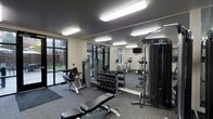 This fitness center is designed to support a full range of workout routines in a functional, well-appointed space. The room features a dark ceiling contrasted against neutral-toned walls, creating a focused, gym-like atmosphere. Overhead, multiple flush-mounted fluorescent light fixtures ensure the space is brightly and evenly lit throughout.

Large black-framed windows line one wall, inviting natural light and offering views of an adjacent outdoor courtyard area, adding an open, airy feel to the interior. The flooring consists of durable grey carpet, providing comfort and traction during workouts.

Strength training is well-represented, with a multi-station Inspire cable machine anchoring one side of the room, a tiered dumbbell rack stocked with a full range of free weights, and multiple adjustable benches for varied exercises. A dedicated curl/preacher bench adds further versatility to the strength equipment selection.

For cardio, upright bikes and a treadmill are available, with a wall-mounted flat-screen television nearby for entertainment. Full-length mirrors along one wall visually expand the space and allow users to monitor their form. Overall, the fitness center offers a clean, modern, and comprehensively equipped environment for effective training.