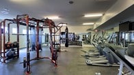 This fitness center offers a comprehensive workout environment designed to accommodate a wide variety of training styles. The space is anchored by a large, multi-functional cable and strength training rig finished in a bold red frame, providing versatile options for resistance exercises. Suspended nearby is a heavy punching bag, ideal for boxing and martial arts training. Along one wall, floor-to-ceiling mirrors visually expand the space while allowing users to monitor their form during workouts. A well-stocked free weight rack lines the mirrored wall, offering a broad selection of dumbbells to suit all fitness levels. On the opposite side, a row of treadmills and elliptical machines provides ample cardio options for members. The flooring throughout is a durable rubber surface, offering both safety and comfort during high-impact activities. Overhead, bright fluorescent lighting ensures the entire space is evenly and thoroughly illuminated. The textured ceiling and clean white walls give the facility a practical, no-frills aesthetic focused entirely on function. Overall, this fitness center is thoughtfully laid out to maximize usability, making it a well-rounded facility suitable for strength training, cardio, and functional fitness routines.