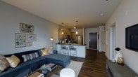 This contemporary single-floor residence showcases a thoughtfully designed open-concept layout that seamlessly connects the living area and kitchen into one cohesive, functional space. Rich, dark hardwood flooring runs throughout, grounding the interior with warmth and sophistication.

The living area is generously proportioned, providing ample room for comfortable seating arrangements. A sectional sofa in deep blue anchors the space, complemented by layered textiles and a classic area rug that adds texture and dimension underfoot.

The kitchen is a standout feature, boasting warm wood cabinetry, dark countertops, and stainless steel appliances. A peninsula with bar seating serves as both a casual dining destination and a natural visual divider between the living and cooking areas. Three pendant lights hang above, casting a warm, inviting glow that enhances the modern aesthetic.

The entry corridor, visible from the main living space, provides a clear sense of flow and is finished in a clean, neutral palette consistent with the rest of the home. The overall ambiance is modern and refined, with a neutral gray and warm wood color scheme that creates a stylish yet welcoming atmosphere throughout.