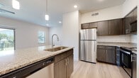 This single-floor unit presents a contemporary, well-appointed interior with a clean, neutral palette throughout. The kitchen serves as a central highlight, featuring a large peninsula island topped with speckled granite countertops, an integrated stainless steel sink with a pull-down faucet, and a stainless steel appliance suite including a refrigerator, electric range, and over-the-range microwave. Dark wood-toned cabinetry with sleek hardware contrasts elegantly against the white subway tile backsplash, while light wood-style plank flooring anchors the space with warmth.

The open-concept layout connects the kitchen seamlessly to the living area, which is generously sized and bathed in natural light from a large sliding glass door that opens to a private balcony with an iron railing. An additional window further enhances brightness. The bedrooms feature plush carpeting, ceiling fans, and ample natural light from windows with blinds. Closet access is provided via paneled interior doors.

The bathroom is cleanly designed with matching granite-topped dark cabinetry, a large vanity mirror, a full tub-shower combination with white tile surround, and light wood-style flooring. Recessed lighting and a well-ventilated layout complete the space, contributing to an overall ambiance that is modern, cohesive, and move-in ready.