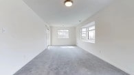 Located at 1 Greenview Circle in West Sayville, NY, this single-floor residence at Greenview West offers a clean, modern living environment designed with comfort and functionality in mind. The interior is defined by crisp white walls and smooth ceilings that lend the space a fresh, airy feel throughout. Plush gray wall-to-wall carpeting runs across the main living area, providing warmth and a soft underfoot experience. Multiple windows along the perimeter flood the space with generous natural light, framing views of the surrounding wooded landscape and creating a bright, welcoming atmosphere. A sliding glass door provides convenient access to the outdoors, further enhancing the connection to the natural surroundings. Flush-mount ceiling fixtures provide consistent overhead lighting, complementing the abundant daylight. A staircase with a warm wood handrail leads down to the entry level, hinting at a well-organized layout that transitions seamlessly between levels. The open layout of the main living area allows for flexible furniture arrangements, making it well-suited for a variety of living styles. The overall design aesthetic is clean and neutral, providing a versatile canvas for personalization.